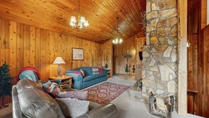 A cozy wooden interior with a stone fireplace, a blue sofa, leather armchairs, and a chandelier. - Cascade Cabin | Deck, 2 Living Rooms + Fireplaces (Cascade)