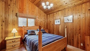 A wooden-paneled bedroom with a bed, bedside table, lamp, and framed pictures on the wall.