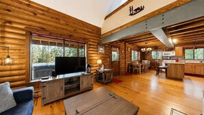 A log cabin interior with a TV, wooden furniture, a dining area, and a kitchen.
