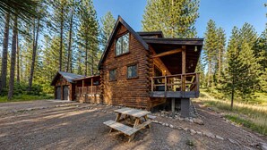 A log cabin with a porch, a covered deck, and a picnic table in a forest setting.