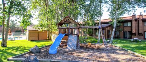 A playground with a slide and climbing area, surrounded by trees and a building in the background.