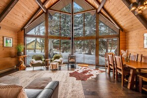 L’intérieur d’une cabane en rondins, avec une grande table à manger en bois, une cheminée et une vue sur un paysage enneigé.