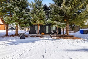 Una cabaña de color negro, rodeada de terreno y árboles cubiertos de nieve.