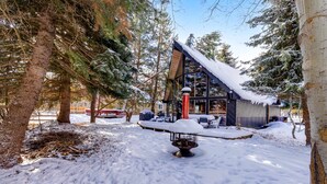 Un espacio exterior cubierto de nieve con una terraza de madera, una fogata y un edificio moderno con ventanas amplias y techo inclinado.