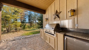 An outdoor kitchen with a built-in grill, a star decoration, and a stone pathway leading to a forested area.