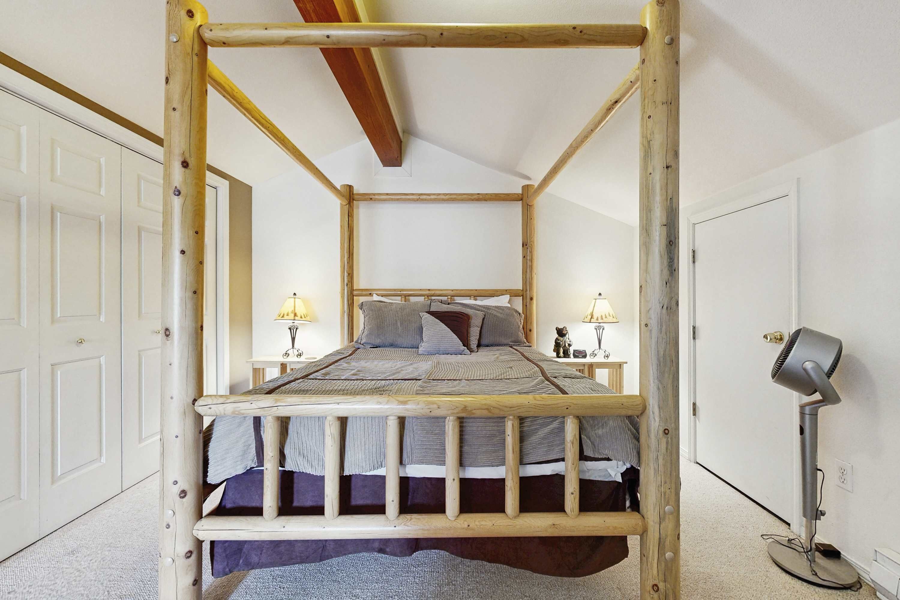 A wooden four-poster bed with a headboard, two bedside lamps, and a floor lamp in a bedroom with white walls and a door on the left.