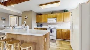 Eine Küche mit Holzausbau, einem weißen Kühlschrank, einer Mikrowelle und einem Spülbecken. An der Kücheninsel stehen drei hölzerne Barhocker.