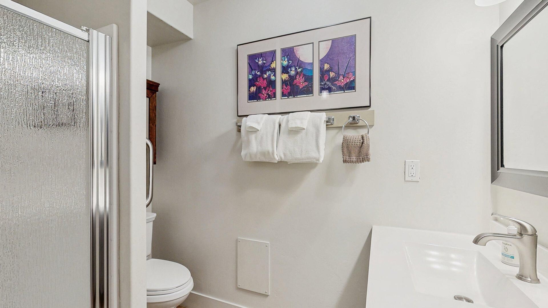 A bathroom with a toilet, a sink, and a shower. There are towels hanging on a rack and a framed picture on the wall.