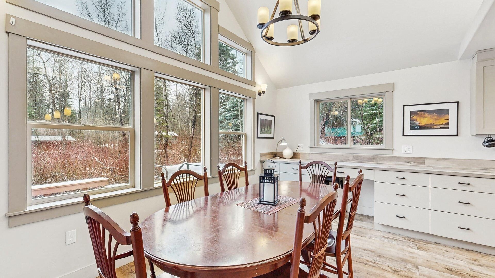 Une salle à manger avec une grande table et des chaises en bois, de grandes fenêtres donnant sur la verdure, une cuisine avec des meubles blancs et un lustre.