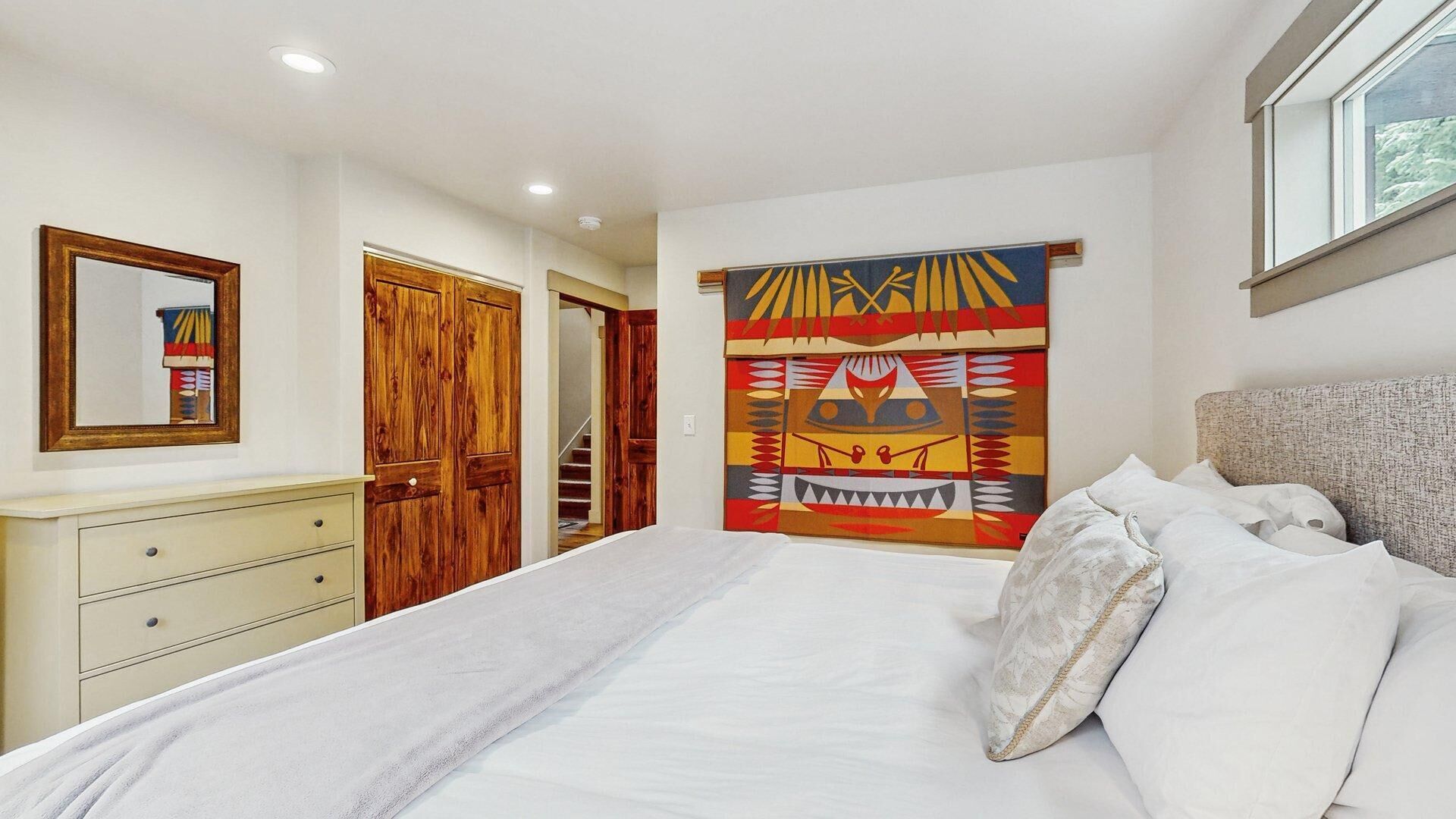 A bedroom with a bed, a dresser, a mirror, a wooden door, and a colorful tapestry.