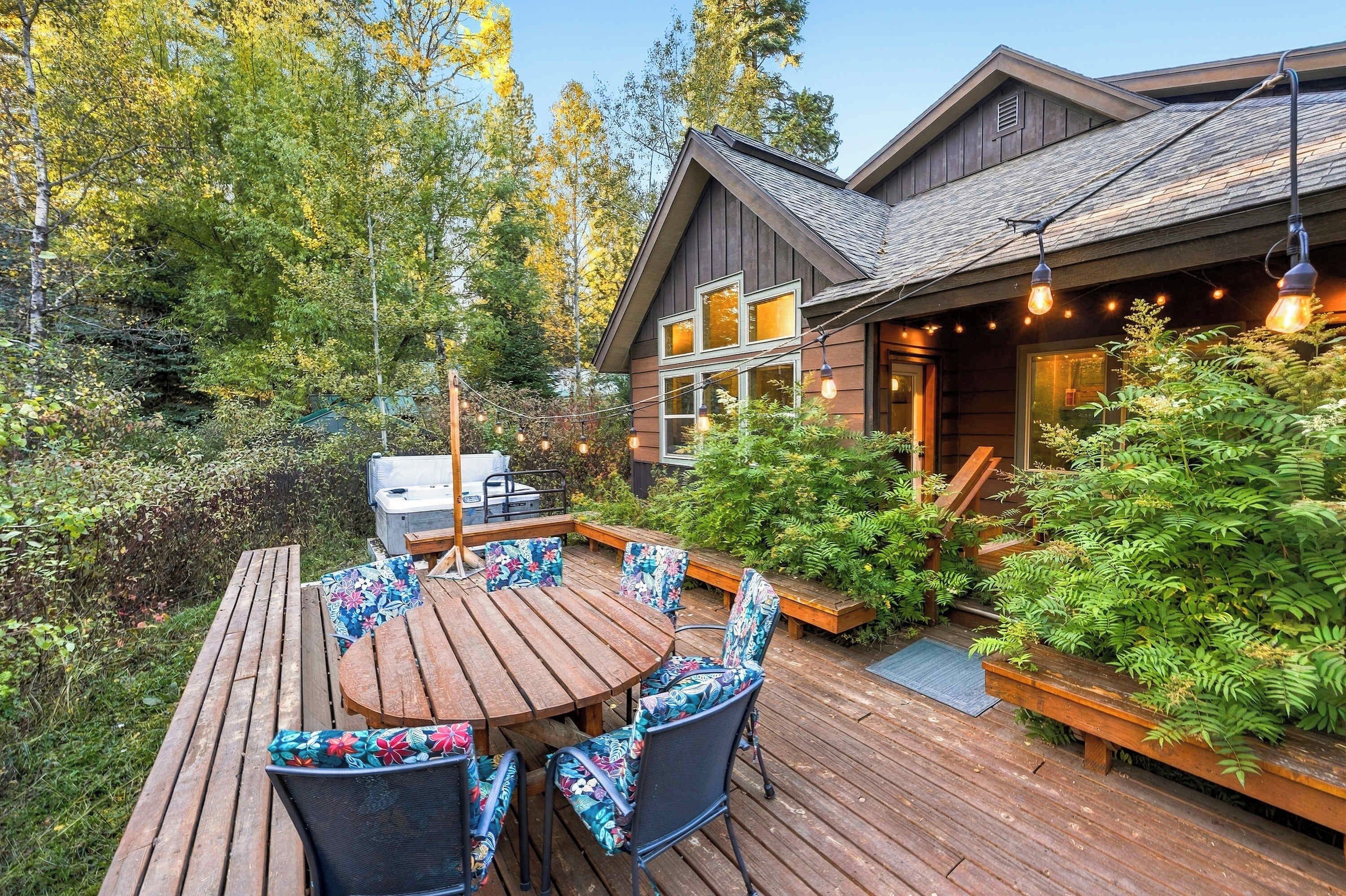 A wooden deck with a round table and chairs, surrounded by lush greenery and a hot tub.