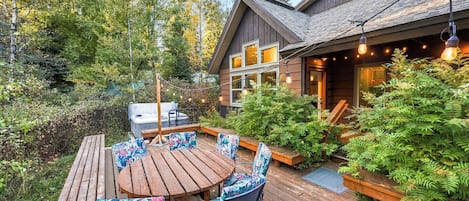 A wooden deck with a round table and chairs, surrounded by lush greenery and a hot tub.
