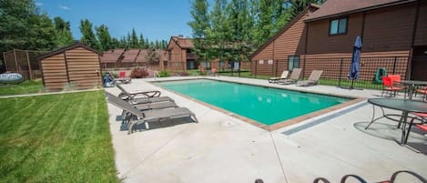 Une piscine rectangulaire, agrémentée de chaises longues et d’une table, entourée d’une cour clôturée et de bâtiments en bois.