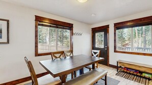 A dining area with a wooden table and chairs, large windows with a view of trees, a door with a coat rack, and a bench with storage. - Downtown McCall Retreat | Wood Stove & Deck (McCall)