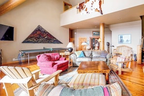 Ein geräumiges Wohnzimmer mit hoher Decke, einem großen Teppich, einem Holztisch und verschiedenen Sitzmöglichkeiten, darunter ein rotes Sofa und zwei Sessel.