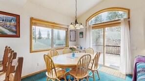 A dining area with a round table, wooden chairs, a vase with flowers, and large windows with curtains. - Spring Mountain Cottage | Pool, Gym, Hot Tub (McCall)