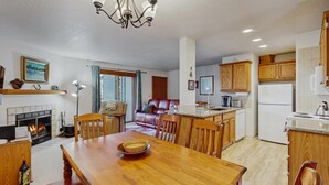 A cozy living room with a fireplace, a dining area with a wooden table, and a kitchen with wooden cabinets and a refrigerator. - Downtown McCall Condo | Fireplace + Balcony (McCall)