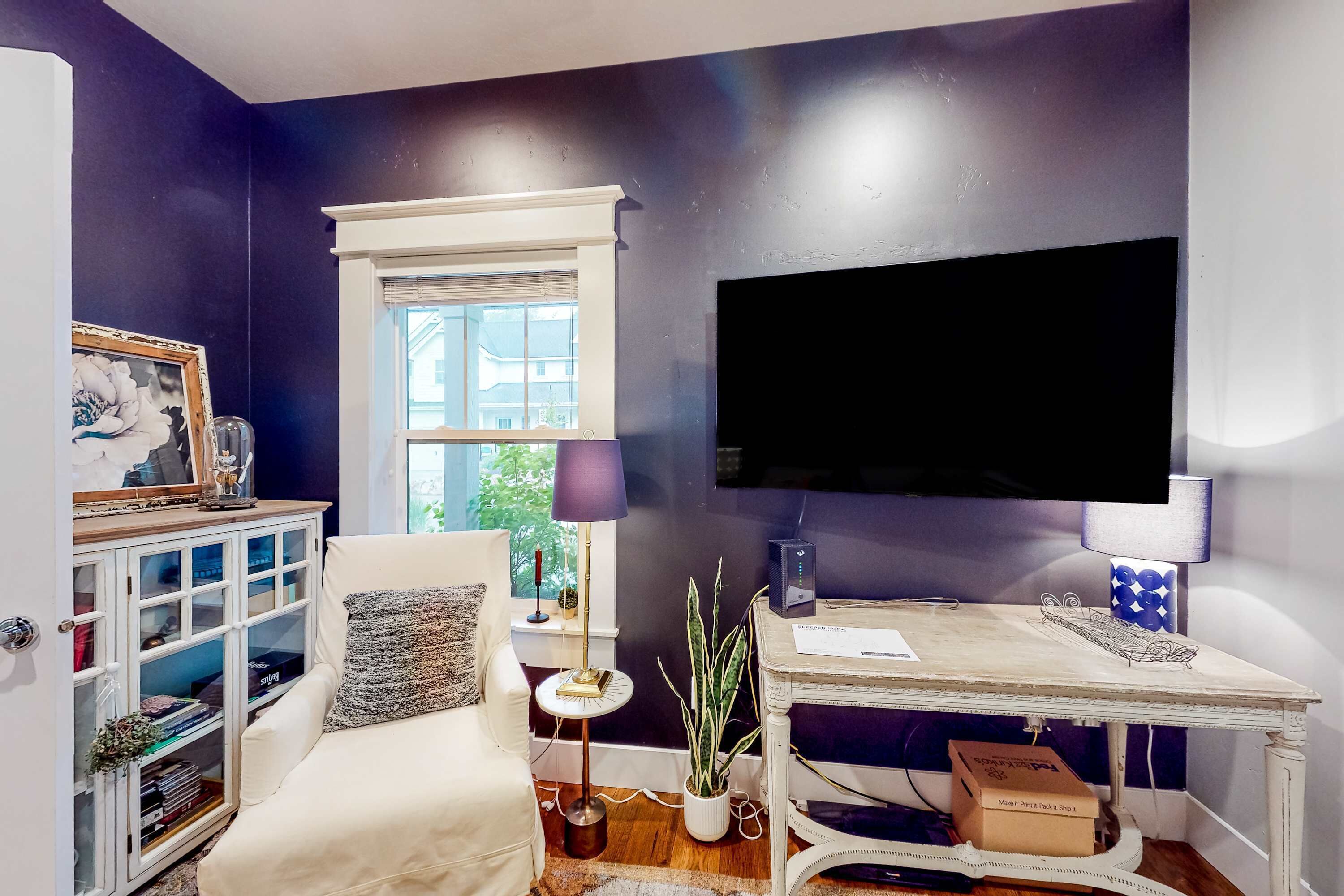 A living room with a white sofa, a small side table, a potted plant, a desk with a lamp, and a television mounted on the wall.