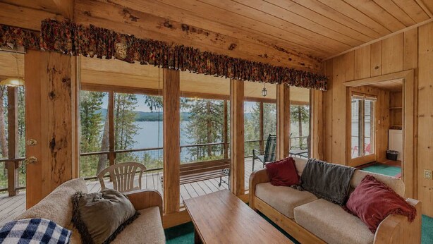 Eine Holzhütte mit einer Couch, einem Tisch und Blick auf einen See durch große Fenster.