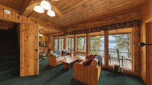 Ein Holzblockhaus-Innenraum mit einer Couch, einem Tisch und Stühlen, große Fenster mit Blick auf Bäume und Wasser.