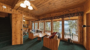A wooden cabin interior with a sofa, table, and chairs, large windows with a view of trees and water. - McCall Lakehouse Private Dock, Beach & Grill (McCall)