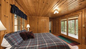 Ein Schlafzimmer einer Holzhütte mit einem Bett, zwei Nachttischlampen, einem Fenster mit Vorhängen und einer Tür, die nach draußen führt.