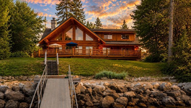 A log cabin with a deck leading to a dock over water, surrounded by a stone wall and trees.