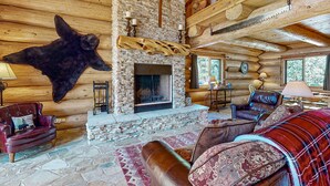 A log cabin interior with a stone fireplace, leather furniture, and a mounted bear skin on the wall.