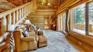 A log cabin interior with a stone floor, wooden staircase, leather furniture, and large windows offering a view of the outdoors.