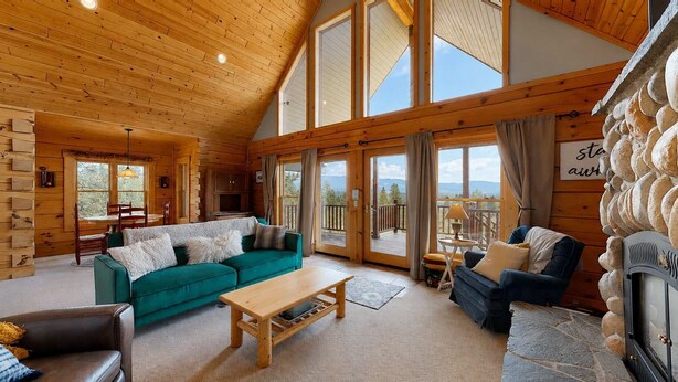 A spacious living area with a wooden ceiling, large windows, a green sofa, a wooden coffee table, and a fireplace. - Mountain View Retreat | Pool & Golf Access (New Meadows)