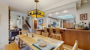 A dining area with a wooden table and chairs, a hanging light fixture, and a kitchen in the background.