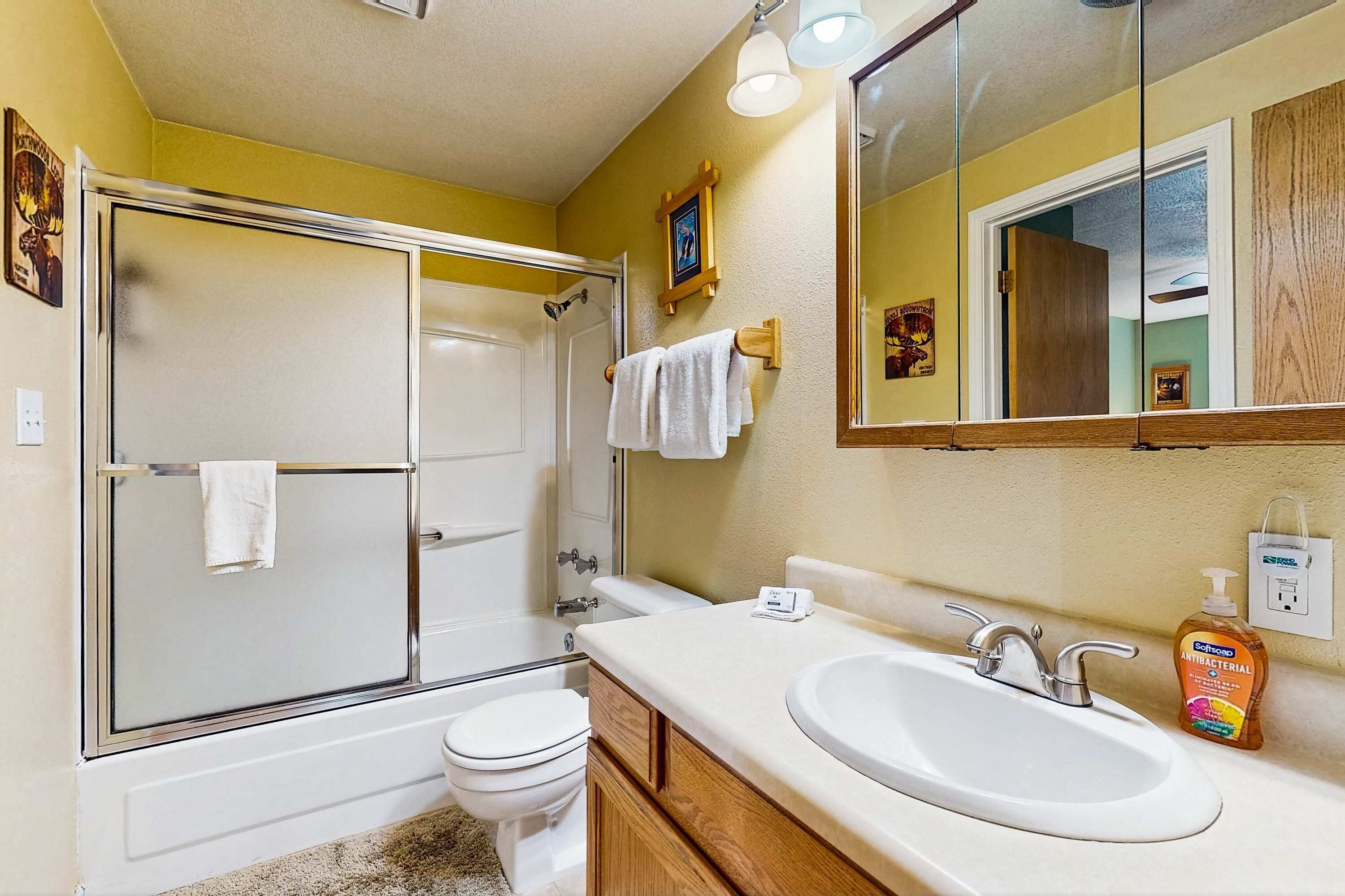 Een badkamer met een douche met glazen wanden, een wit toilet, een wastafel met spiegel erboven en een fles handzeep op het aanrecht.