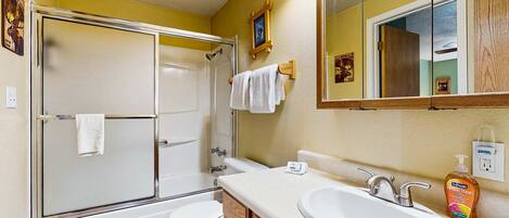 Een badkamer met een douche met glazen wanden, een wit toilet, een wastafel met spiegel erboven en een fles handzeep op het aanrecht.