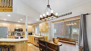 A modern kitchen with wooden cabinets, a central island with stools, a dining table with chairs, and a chandelier.