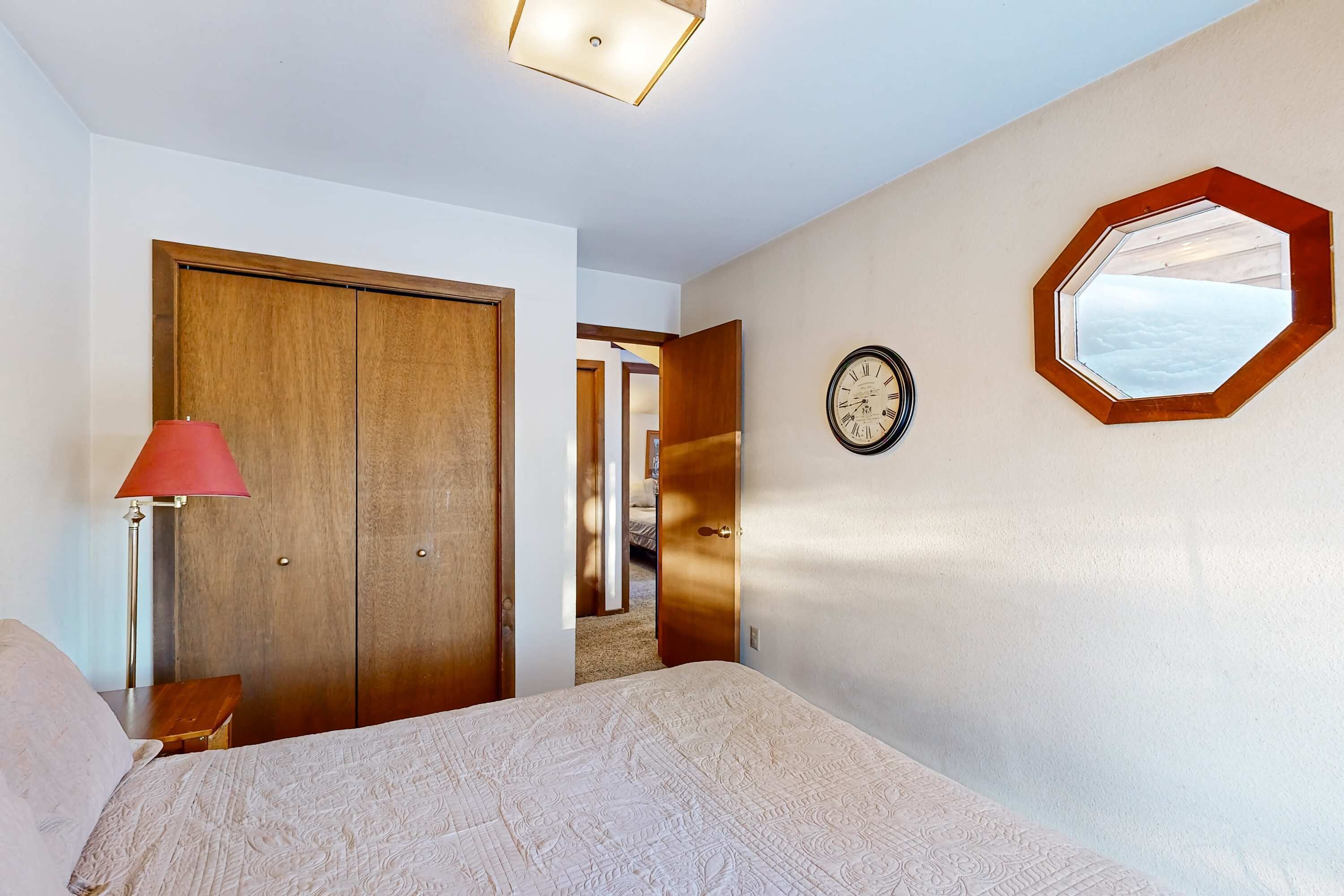 A bedroom with a bed, a wooden wardrobe, a red lamp, a clock, and a hexagonal mirror on the wall.