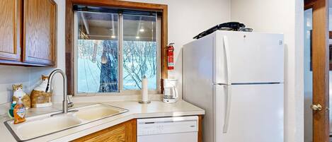 Une cuisine avec des meubles en bois, un lave-vaisselle blanc, un réfrigérateur blanc, une fenêtre donnant sur des arbres et un extincteur.