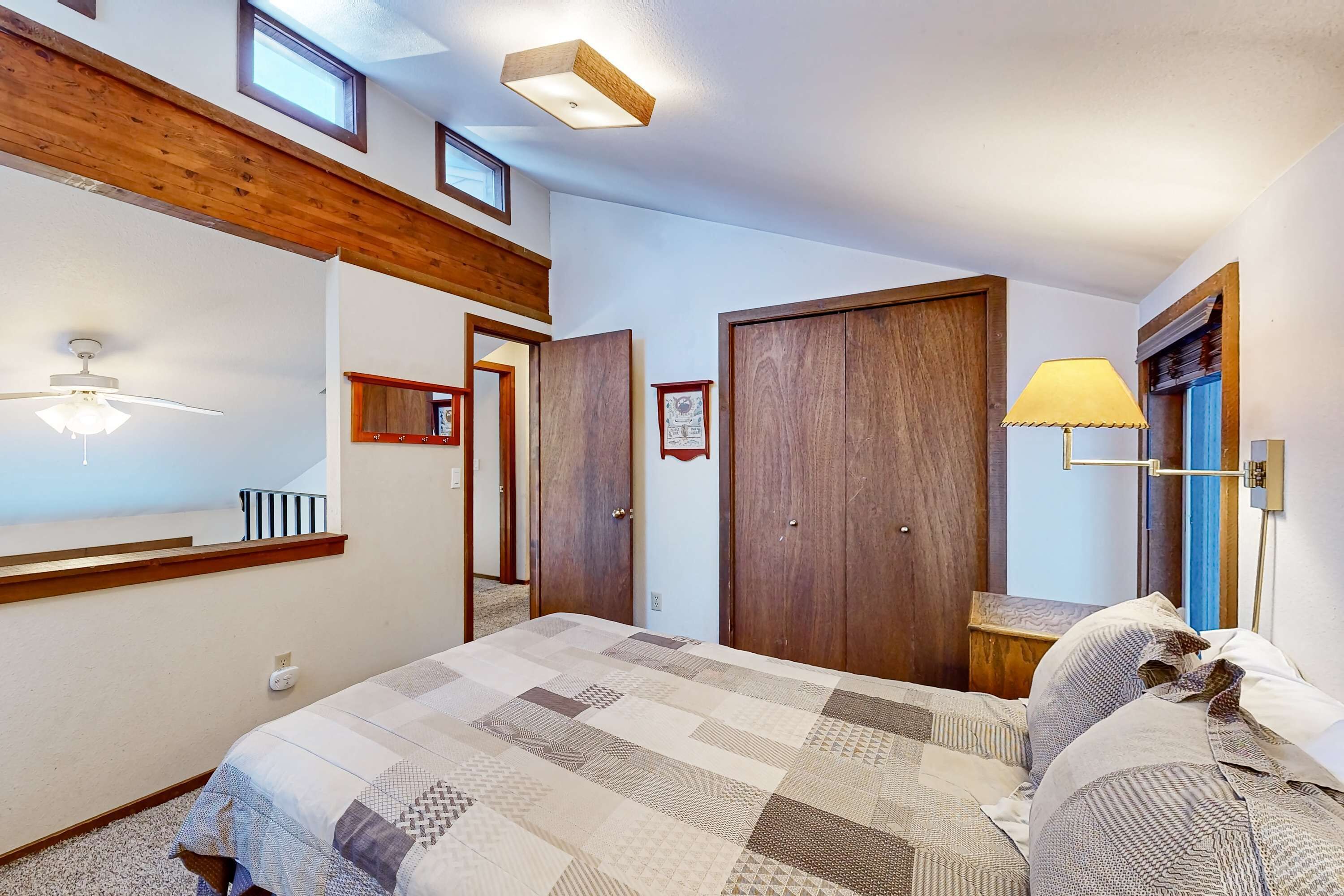 A bedroom with a large bed, wooden ceiling beams, a ceiling fan, a nightstand, a lamp, and a door with a picture frame.