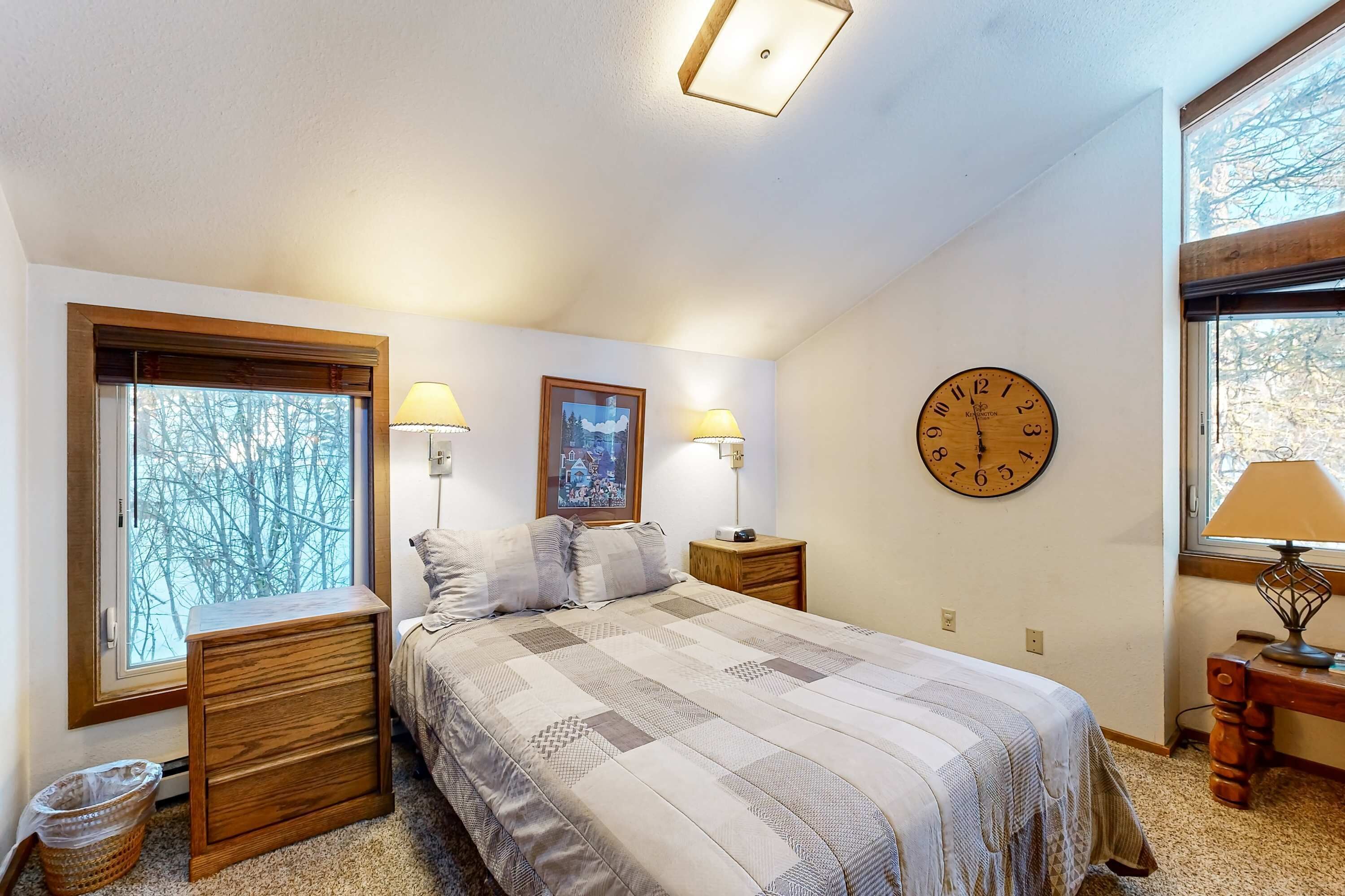 A bedroom with a large bed, two bedside tables with lamps, a wall clock, and a window with a view of trees.