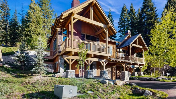 Une grande cabane en rondins à plusieurs étages, dotée d’un balcon, entourée d’arbres et bordée d’un sentier en pierre.