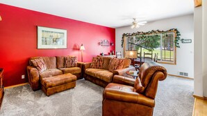 A living room with a sectional sofa, a leather armchair, and a coffee table. There is a window with a view of a deck and trees.