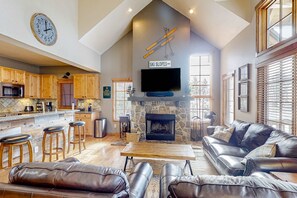 A spacious living area with a stone fireplace, leather sofas, a wooden coffee table, and a kitchen with wooden cabinets and a built-in TV. - Tamarack Chalet, Hot Tub, Ski-In/Out, Sleeps 8 (Donnelly)