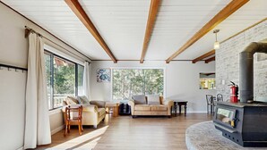 Ein gemütliches Wohnzimmer mit Kamin, Holzbalken, einer Couch und Blick auf Bäume durch große Fenster.