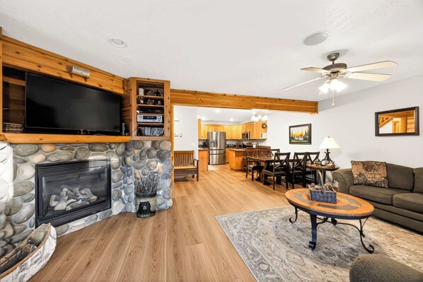 A spacious living room with a stone fireplace, a flat-screen TV mounted on a wooden wall, a dining area with a wooden table and chairs, and a kitchen in the background. - McCall 3 Bedroom Condo | Walk to Lake, Park & Golf (McCall)