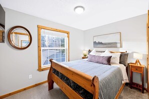 Une chambre à coucher avec un lit en bois, une fenêtre, une table de chevet, une commode, une lampe, un miroir et un tableau accroché au mur.