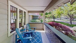 A covered porch with two blue Adirondack chairs, a patterned rug, and a view of a landscaped garden with trees and shrubs. - Historic Craftsman Yard, Patio & Fireplace (Boise)