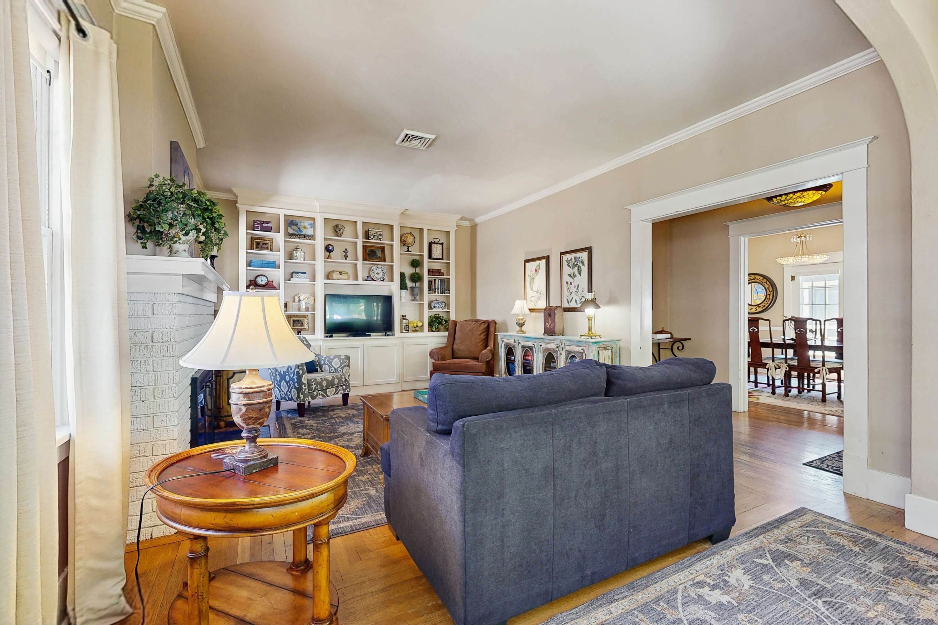 A living room with a grey sofa, a wooden side table with a lamp, a TV, and a bookshelf filled with various items.