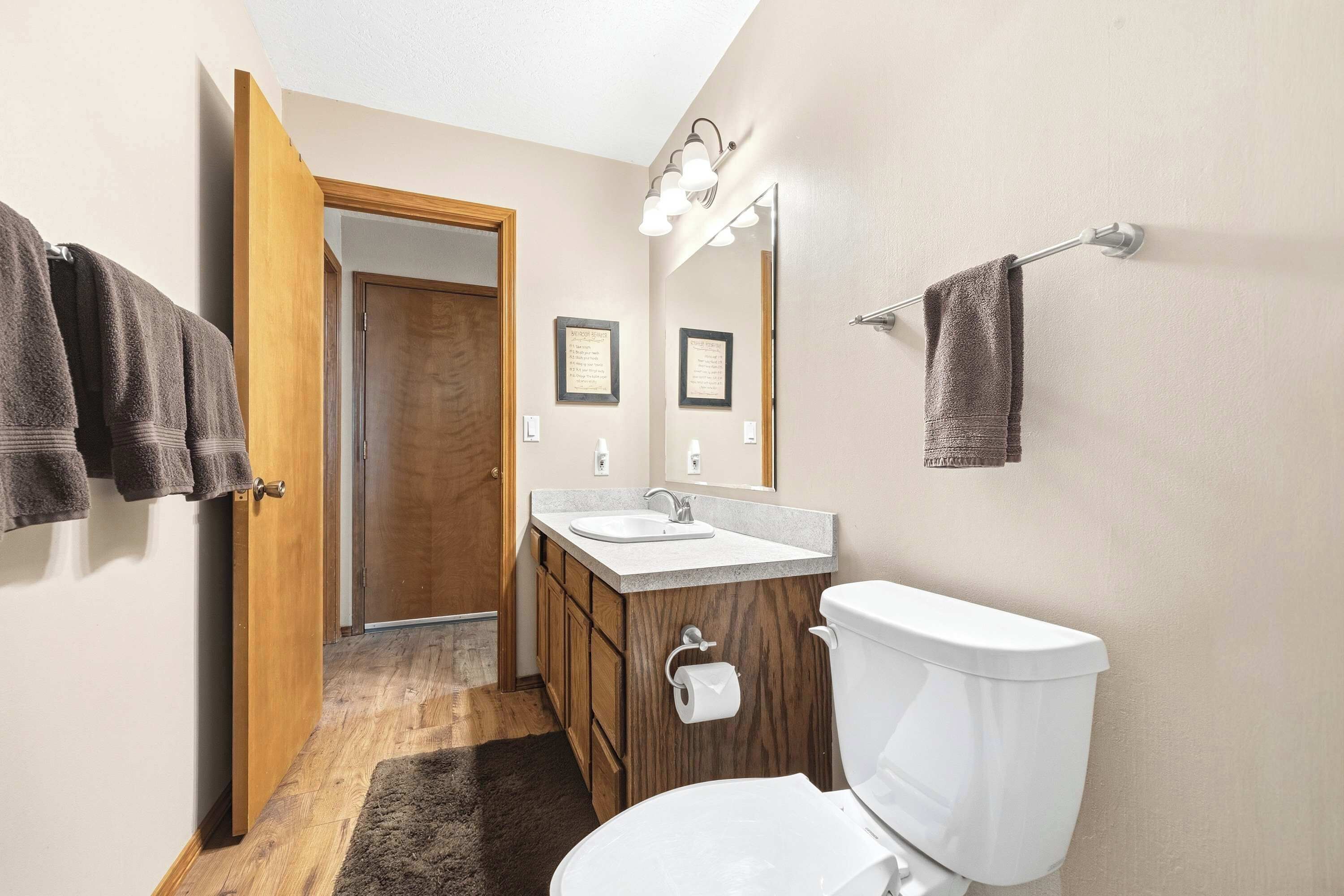 Ein Badezimmer mit Toilette, Waschbecken und Holzschränken. Handtücher hängen an einem Wäscheständer und eine Tür führt in einen anderen Raum.