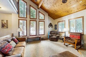 A cozy living room with a wooden ceiling, large windows, a sofa, a rocking chair, and a bench.