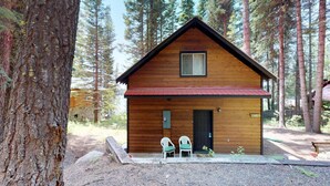 A wooden cabin with a red roof, a small porch with two chairs, and a stone wall.