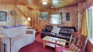 A wooden cabin interior with a sofa, a chair, a table, and a window with curtains.